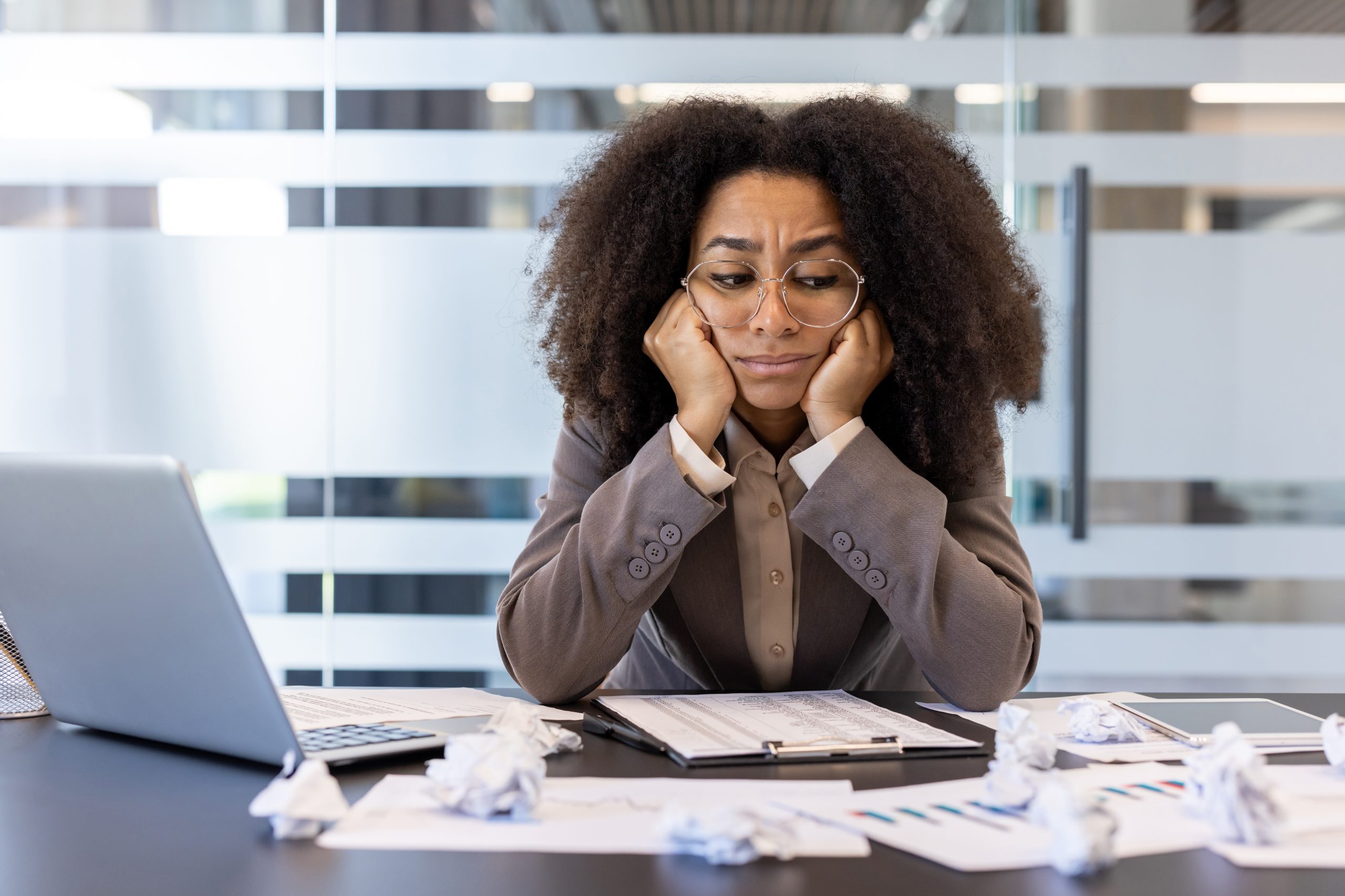 A frustrated Black woman CEO sits at her desk wiht a laptop and crumpled notepaper around her.
